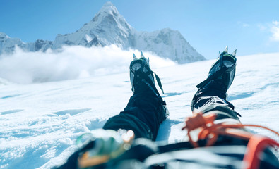 POV-Shooting der Lags eines Höhenbergsteigers in Steigeisen. Er liegt und ruht auf einem Schnee-Eisfeld mit Ama Dablam (6812 m) Gipfel, der mit Wolken bedeckt ist. Urlaubskonzept für extreme Menschen