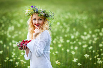 Young beautiful blond woman in white dress and floral wreath standing and eating strawberry on summer day