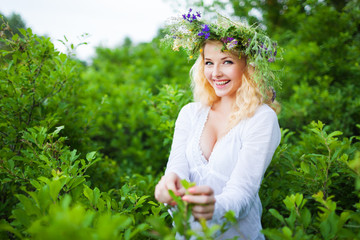 Young beautiful blond woman in white dress and floral wreath standing and smiling on summer day