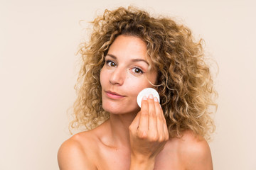 Young blonde woman with curly hair removing makeup from her face with cotton pad
