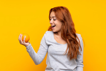Teenager redhead girl holding an orange over isolated yellow background