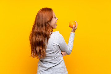 Teenager redhead girl holding an orange over isolated yellow background