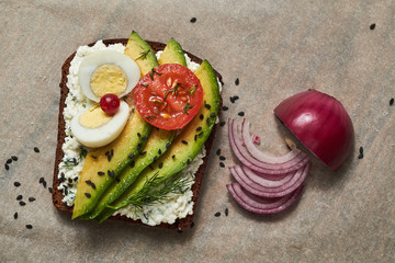 Healthy toast with cottage cheese, avocado, egg and tomato. Nearby lies a purple chopped onion and black sesame seeds. Healthy eating concept.
