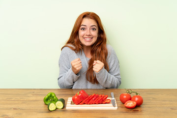 Teenager redhead girl with vegetables in a table celebrating a victory