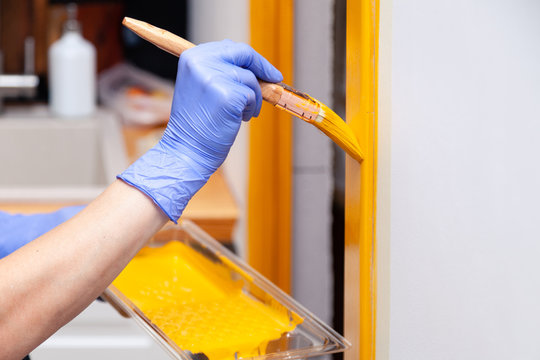 Closeup Woman Hand In Purple Rubber Glove With Paint Brush Painting Natural Wooden Door With Yellow Paint, Creative Design House Renovation Theme. How To Paint Wooden Surface. Selected Focus