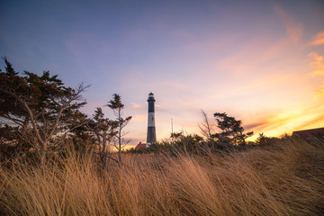 Golden sunset light illuminating beach grass in front of a stone lighthouse, with wispy clouds overhead in the sky. Fire Island National Seashore, New York. 