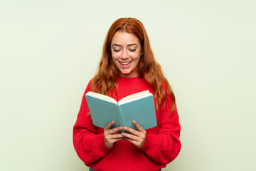 Teenager redhead girl with sweater over isolated green background holding and reading a book