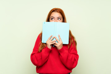 Teenager redhead girl with sweater over isolated green background holding and reading a book