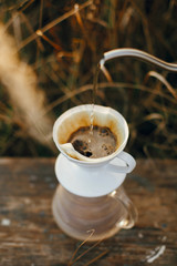 Hand pouring hot water from steel kettle in glass flask dripper with filter on background of rural countryside, herbs in sunset. Alternative coffee brewing. Atmospheric rustic moment. Vertical image