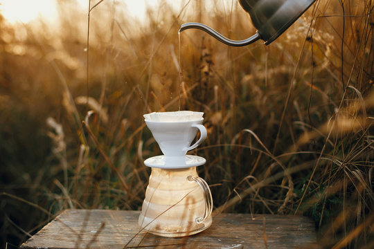 Hand pouring hot water from steel kettle in glass flask dripper with filter on background of rural countryside among herbs in sunset. Atmospheric rustic moment. Alternative coffee brewing - Powered by Adobe
