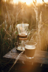 Hot coffee in glass cup and glass flask on background in sunny warm light in rural herbs. Alternative coffee brewing outdoors in travel. Atmospheric rustic tranquil moment. Vertical image