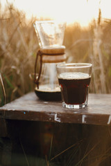 Alternative coffee brewing outdoors in travel. Hot coffee in glass cup and glass flask on background in sunny warm light in rural herbs. Atmospheric rustic tranquil moment. Vertical image