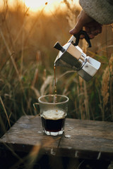 Hipster pouring fresh hot coffee from geyser coffee maker into glass cup in sunny warm light in...