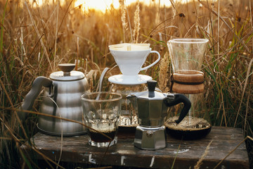 Alternative coffee brewing outdoors in travel. Steel kettle, hot coffee in cup, coffee dripper,  geyser maker, glass flask with filter on background of sunny warm light in rural herbs.