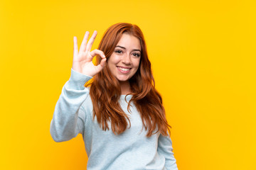 Teenager redhead girl over isolated yellow background showing ok sign with fingers