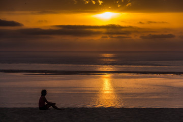 Side view of a young woman seating on the top of the Grande Dune du Pilat and seeing on the Atlantic Ocean and yellow sunset