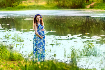 Beautiful pregnant woman mother to be in wreath and blue dress standing on bank of scenic river in sunset time in summer. Pregnancy and healthy organic close to nature lifestyle.