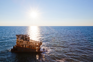 Fototapeta na wymiar Sunken rusty cargo ship in still blue sea waters with bright sun over water horizon on summer clear day with blue sky. Still landscapes and abandoned places concept