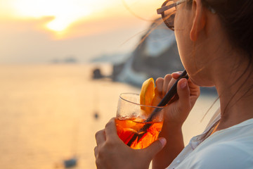 Happy young woman with orange cocktail in glass drink at sunset in front of the sea on Ponza island coast, on a wall with view of the ocean. Smiling at camera.