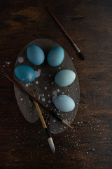 Dark Easter concept. Paint brushes and painted blue eggs with light dots on the artist's palette. Dark photo. Dark and light. Wooden background