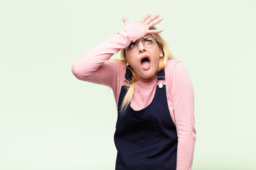 young pretty blonde woman raising palm to forehead thinking oops, after making a stupid mistake or...