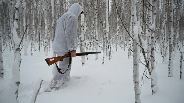 A hunter in the winter in the woods in a white camouflage suit hunts a hare.