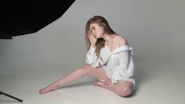 behind the scenes on photo shoot: beautiful young model wearing shirt posing for photographer near softbox on white background, flash lights. model tests of caucasian teen with bare feet in studio