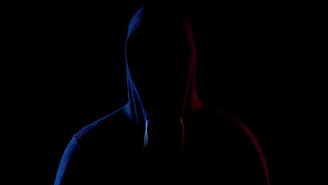 A young man is standing in the hood hiding his face. An attacker is standing in the dark. Blue and red light falls on the person on a black background.