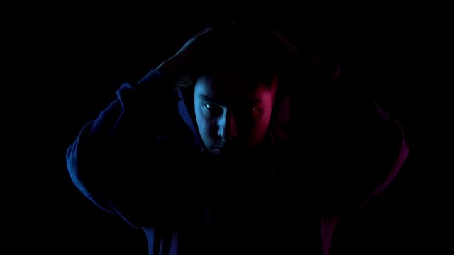 A young man takes off his hood. An attacker is standing in the dark. Blue and red light falls on the person on a black background.