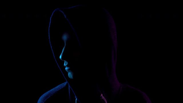 A young woman in a hood looks around. An attacker is standing in the dark. Blue and red light falls on a person on a black background.