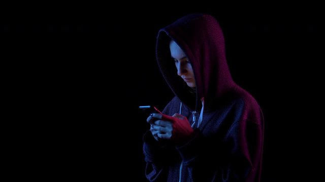 A young woman in a hood with a phone in her hands. Hacker makes a hack through the phone. Blue and red light falls on a person on a black background.