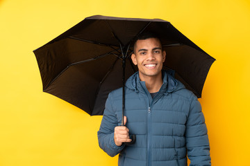 Young handsome man holding an umbrella over isolated yellow background smiling a lot © luismolinero