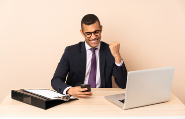 Young business man in his office with a laptop and other documents with phone in victory position