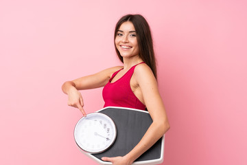 Pretty young girl with weighing machine over isolated pink background holding a weighing machine...