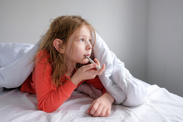 little girl in sleepwear on a white bed sits with a thermometer and measures the temperature