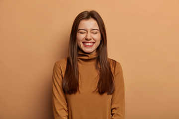 Pretty amused woman has long dark straight hair, laughs from joy, wears brown neck sweater, smirks...
