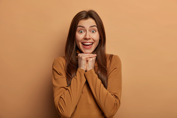 Excited cheerful woman giggles happily, keeps hands near chin, has interesting conversation,...