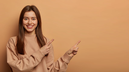 Look, advertise here! Dark haired pretty woman with appealing smile gives positive attitude towards good deal, expresses her recommendation while pointing on right side, wears brown sweater.