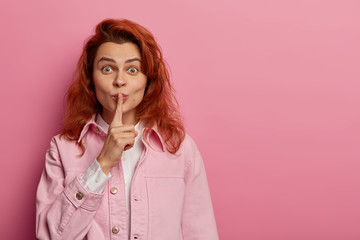 Surprised redhead young woman makes silence gesture, asks keep silence, tells secret information to interlocutor, wears pink jacket, stands indoor. Shh sign. Body language and secrecy concept