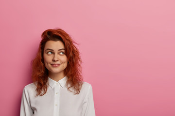 Fototapeta premium Contemplative woman with happy dreamy expression, concentrated aside, thinks about vacation after work, dressed in elegant white shirt, has ginger hair, poses against pink backgroun, makes up idea