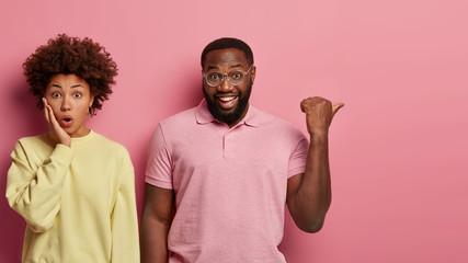 Studio shot of young ethnic couple stand against pink studio background, express various emotions. Shocked Afro American woman gasps from wonder, cheerful bearded man points thumb on free space