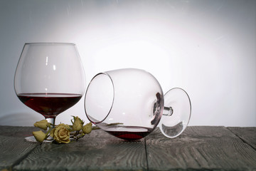 Glasses with red dry wine. One of them lies on its side with the remains of wine. Dried rose flowers. Stand on wooden boards. Shot in backlight.