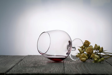 Glasses with red dry wine. One of them lies on its side with the remains of wine. Dried rose flowers. Stand on wooden boards. Shot in backlight.