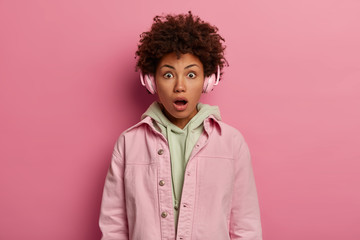 Obraz na płótnie Canvas Beautiful impressed young woman has curly hair listens music with headphones, shocked by terrified relevation, wears pastel jacket, keeps mouth opened, cannot believe eyes. Emotions, lifestyle