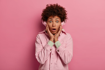 Fototapeta na wymiar Stylish curly haired young woman keeps hands on cheeks, wears pink jacket, listens radio online, uses wireless headphones, has shocked face expression, isolated over rosy wall, likes playlist