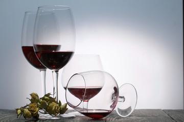 Glasses with red dry wine. One of them lies on its side with the remains of wine. Dried rose flowers. Stand on wooden boards. Shot in backlight.