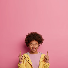 Positive Afro woman points above with closed eyes, feels glad to participate in advertising campaign, has toothy gentle smile, wears yelllow jacket, attracts attention upwards. Your promo content here