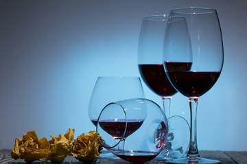 Glasses with red dry wine. One of them lies on its side with the remains of wine. Dried rose flowers. Stand on wooden boards. Shot in backlight.
