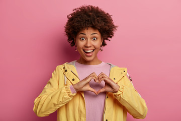 Portrait of beautiful woman with curly hairstyle makes heart gesture over chest, expresses love, says be my valentine, has positive expression, wears yellow anorak, adores boyfriend. Hand sign - Powered by Adobe