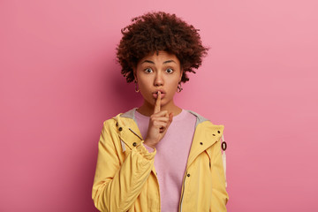 Fototapeta na wymiar Photo of Afro American woman keeps index finger on lips, tells secret information, gossips about colleague, makes hush gesture, wears yellow anorak, has mysterious expression, stands indoor.